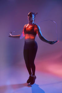 Full body of slim young female in sportswear skipping rope in studio illuminated with purple neon light