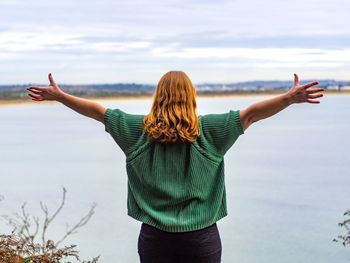 Rear view of woman with arms outstretched standing by sea against sky