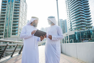 Male coworkers discussing over digital tablet while standing on footbridge in city