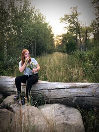 Surprised woman holding flower bouquet while sitting on wood against trees