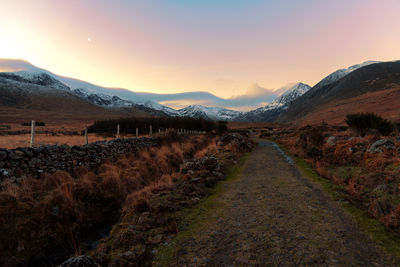 Dirt road amidst field against sky during sunset