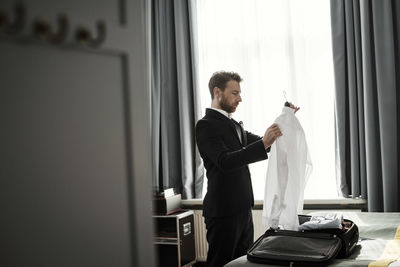 Side view of businessman holding white shirt in coathanger at hotel room