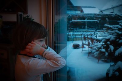 Side view of bored boy looking through window at home during winter