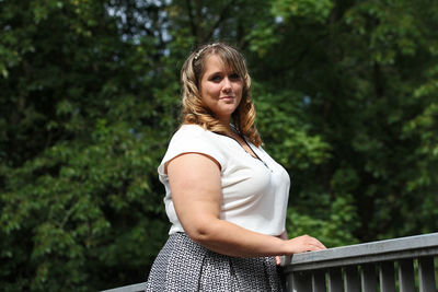 Portrait of young woman standing by railing against trees