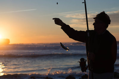 Silhouette man fishing at seashore against sky during sunset