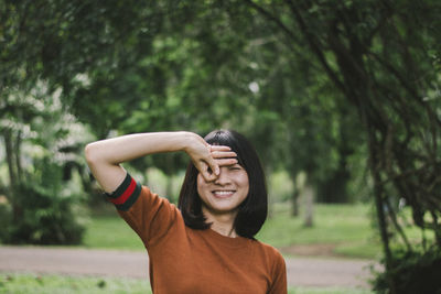 Portrait of smiling young woman gesturing while standing against trees in park