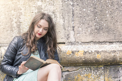 Beautiful young woman reading book while sitting by wall