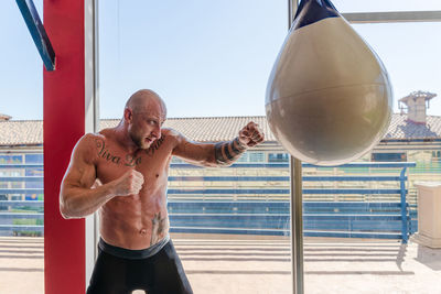 Side view of determined male boxer in boxing gloves punching heavy bag during intense workout in gym