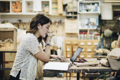 Side view of female upholstery worker using laptop while talking on phone at workbench in workshop