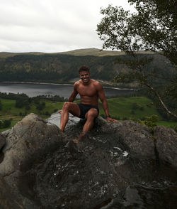 Portrait of shirtless man sitting on rock by lake against sky