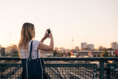 Young woman photographing through mobile phone while standing on bridge against clear sky during sunset