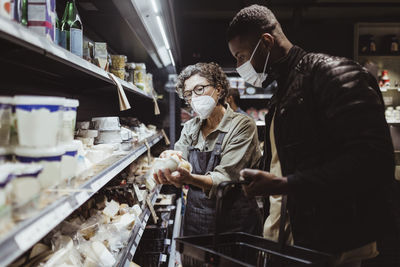 Female owner holding product while male customer standing by in deli store during covid-19