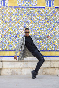 Full body of male in stylish sunglasses standing on tiptoes near colorful ornamental wall in city on summer day