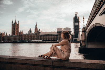 Woman sitting on retaining wall by bridge in city against sky