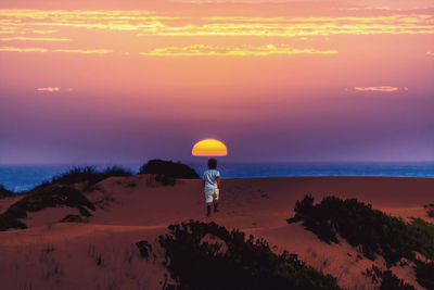 Rear view of boy walking on sand dunes at beach against sky during sunset