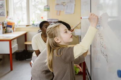 Blond girl solving maths problem while writing on whiteboard in classroom
