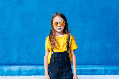 Confident teenager standing with trendy sunglasses blue background and looking at camera