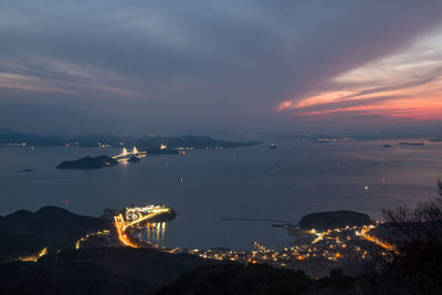 Illuminated city by sea against cloudy sky during sunset