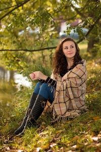 Side view of thoughtful young woman with shawl sitting at grassy lakeshore