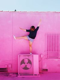 Woman exercising on air conditioner against pink wall during sunny day