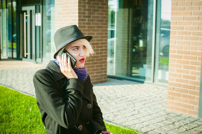 Young woman talking on mobile phone while smoking by built structure