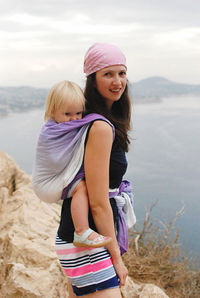 Portrait of young woman holding baby on back standing against sky