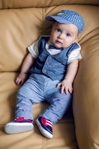 Gay boy kid blonde in a cap , pants, tie and vest sits on a light leather sofa