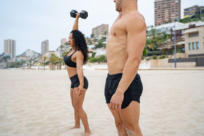 Side view of muscular multiethnic sportspeople exercising with dumbbells while training on sandy beach in summer