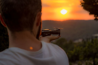 Rear view of man holding camera against sky during sunset