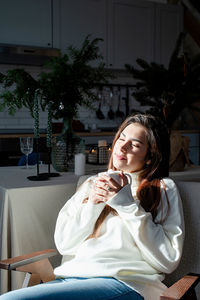 Woman in warm white winter sweater sitting on the window at home at christmas eve holding cup