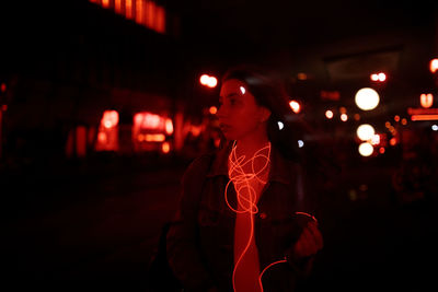 Young woman looking away while standing on footpath at night