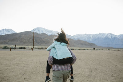 Rear view of father piggybacking daughter while walking on field against clear sky during sunset