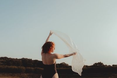 Rear view of young woman holding plastic while standing on land against clear sky