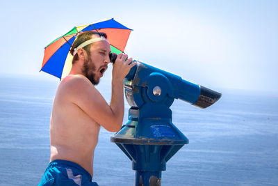 Side view of mid adult man with umbrella looking through coin-operated binoculars at beach