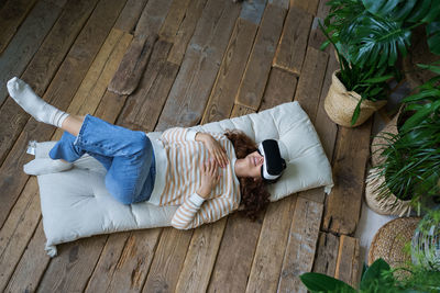 Relaxed caucasian young girl in virtual reality glasses resting on cozy soft mattress in greenhouse.