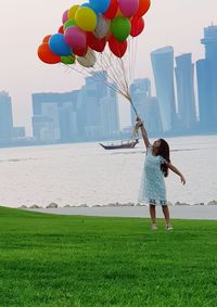 Full length of girl holding colorful helium balloons while standing on grassy field against river and cityscape