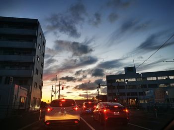 Traffic on road by buildings against sky during sunset