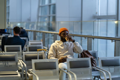 Young african man drinking coffee and eating sandwich while waiting for flying at airport terminal