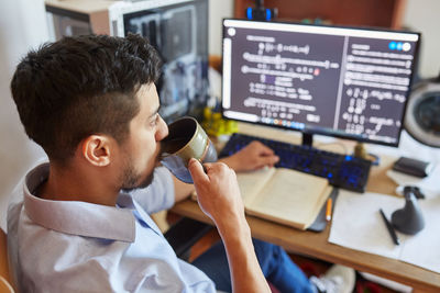 High angle side view of young male student sitting at table with cup of coffee and reading information on computer monitor while preparing for exam in university