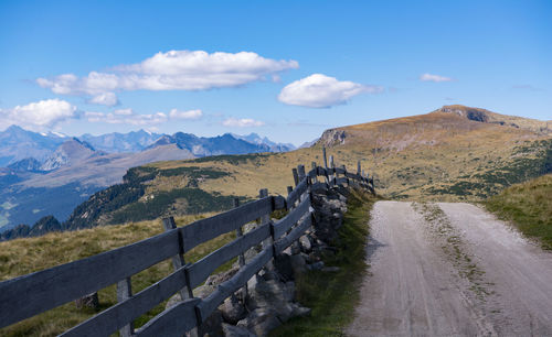 Road by fence leading towards mountain against blue sky