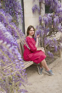 Young woman in red dress sits on a bench in the thickets of blooming wisteria