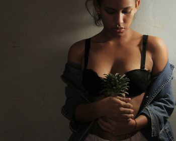 Seductive woman holding pineapple while standing against wall at home