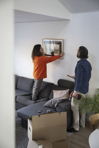 Woman hanging picture frame on wall by man in living room