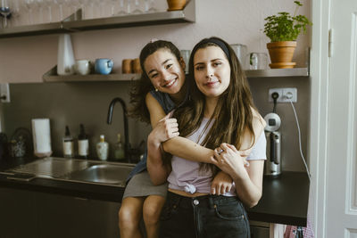 Portrait of happy woman hugging female friend from behind while sitting in kitchen at home