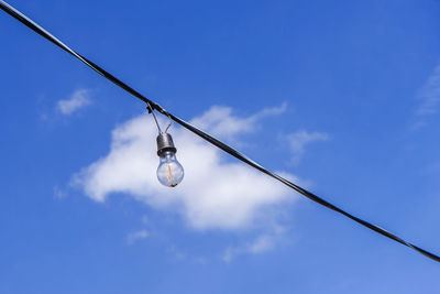 Low angle view of light bulb hanging on cable against blue sky