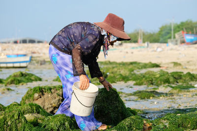 Side view of senior woman holding bucket while working on mossy rocks at beach