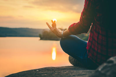 Low section of woman meditating on lakeshore during sunset