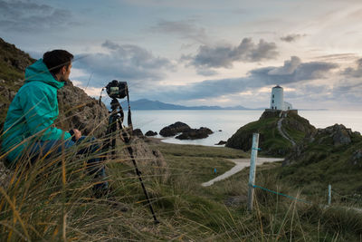 Man sitting by camera on tripod while looking at lighthouse against sky