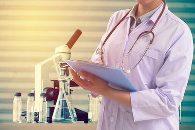 Midsection of female doctor holding on clipboard with chemicals in laboratory