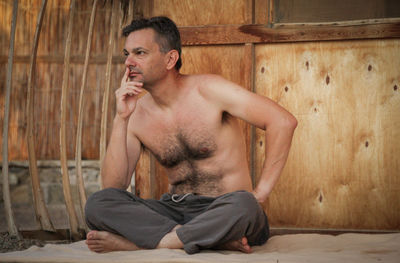 Shirtless man sitting against wooden wall